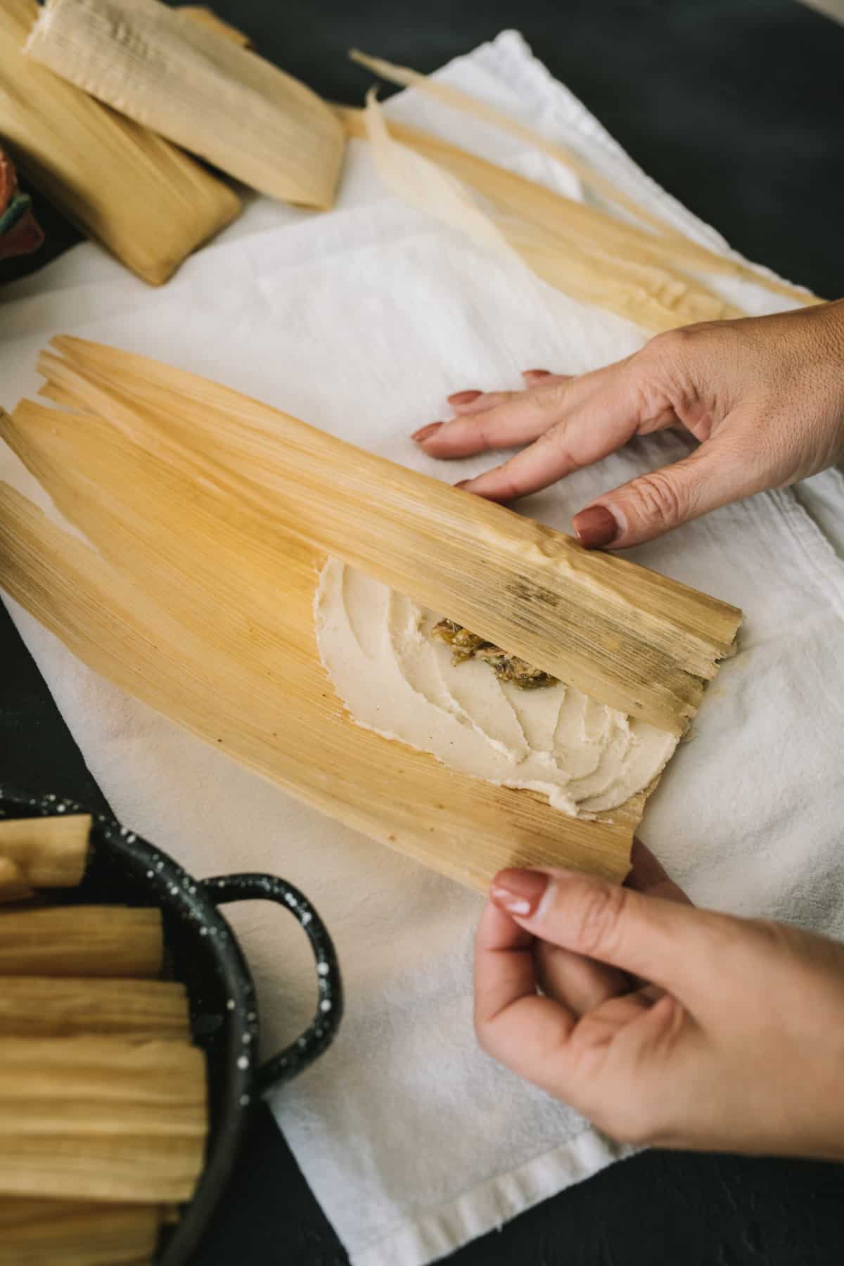 woman's hands rolling in one side of a corn husk to wrap a homemade tamale.