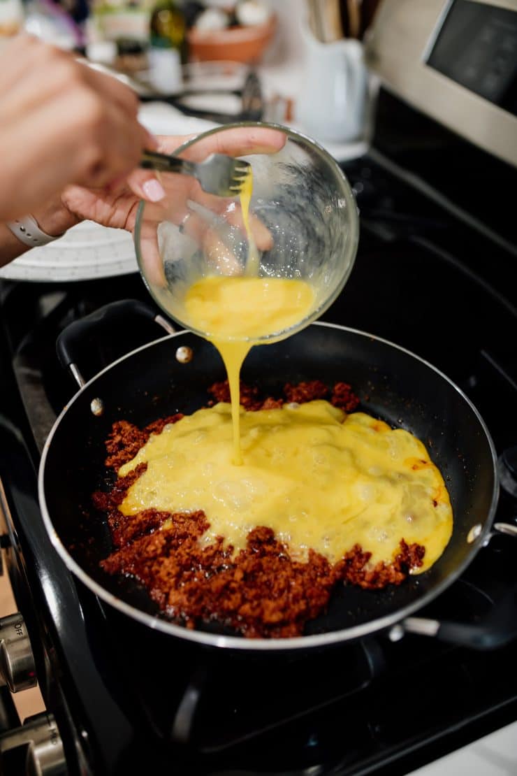 hand pouring beaten eggs into pan with browned chorizo.