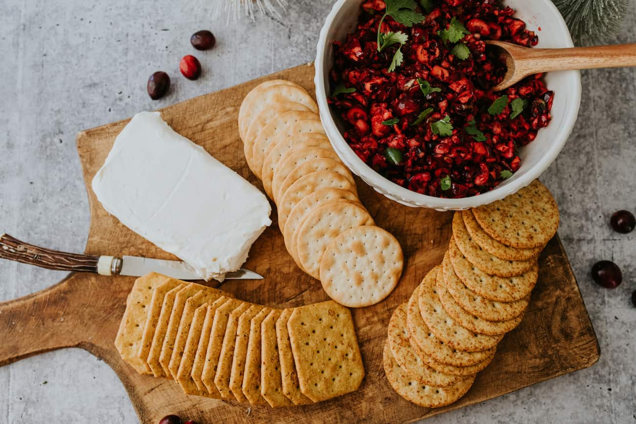 overhead shot of a festive party platter filled with a block of cream cheese, multiple types of crackers, and a white bowl of spicy sweet cranberry jalapeño dip.