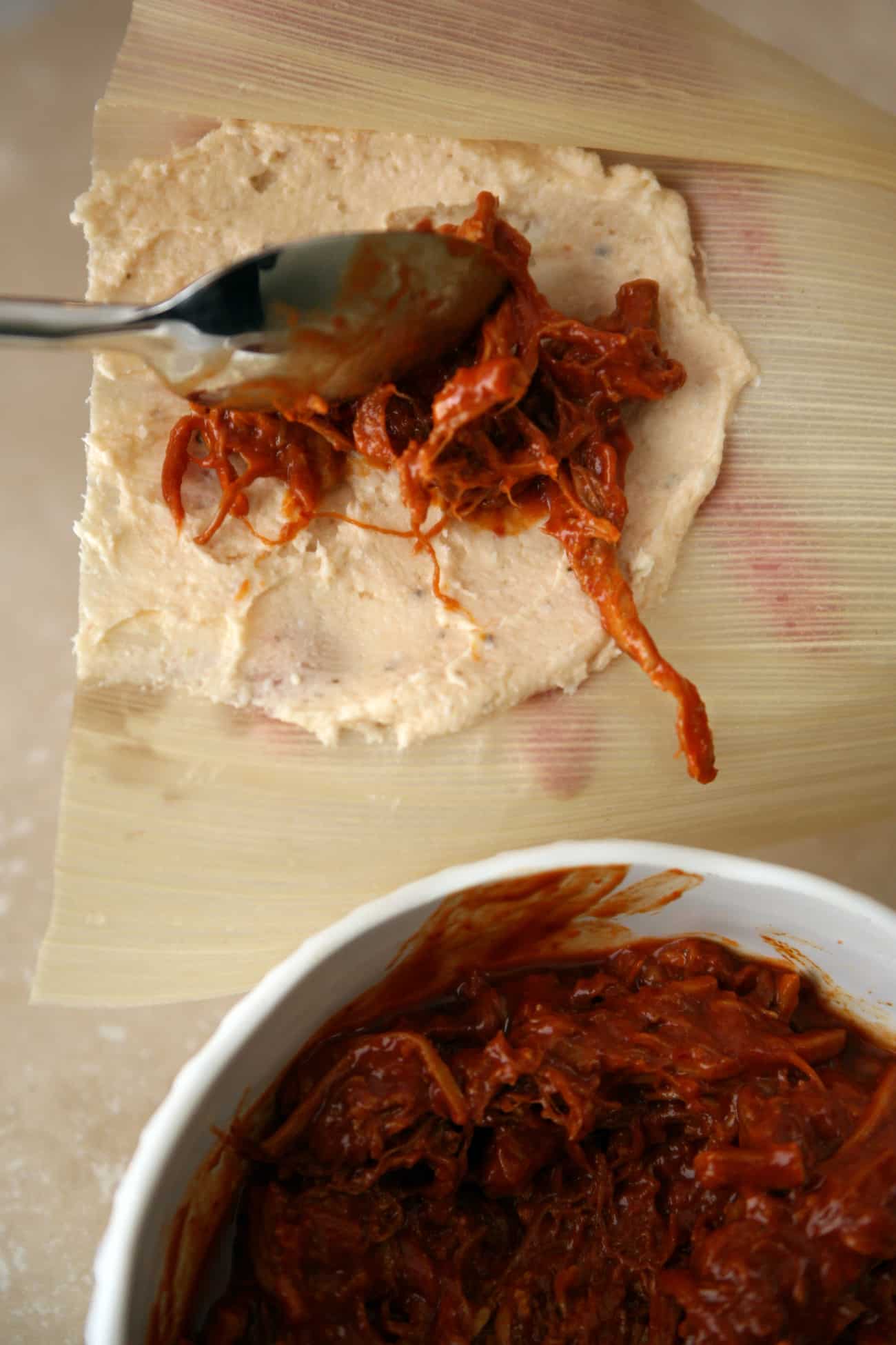 placing a spoonful of red chile pork filling into the center of the masa for rolling into a tamale.