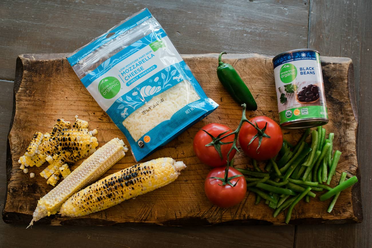 An overhead shot of the ingredients for my Carne Asada Taco Pizza: roasted corn, shredded mozzarella cheese, tomatoes on the vine, a jalapeño for spice, sliced bell peppers and a can of black beans.