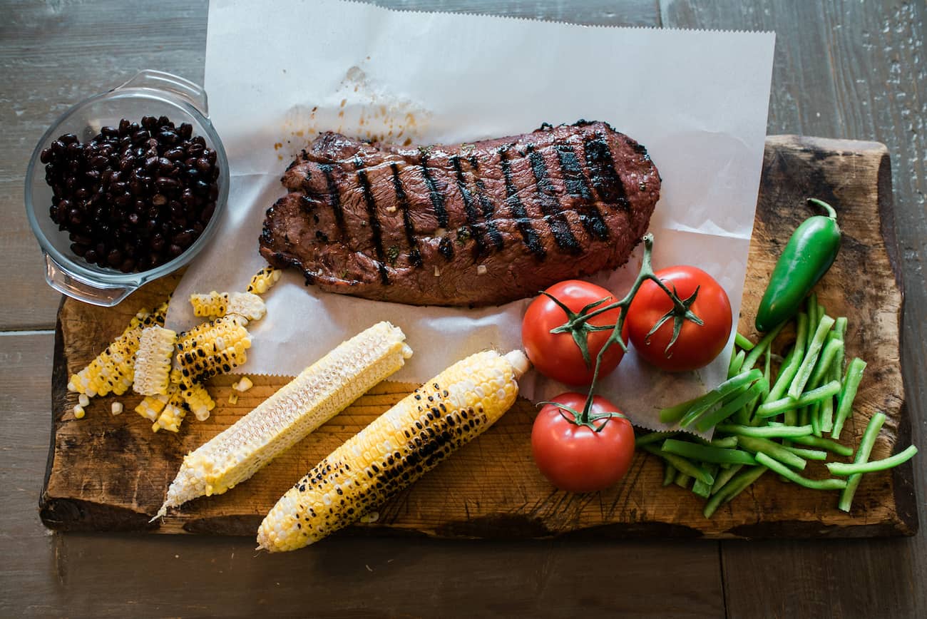 Overhead shot of grilled carne asada, roasted corn and tomatoes on the vine for making Taco Pizza.