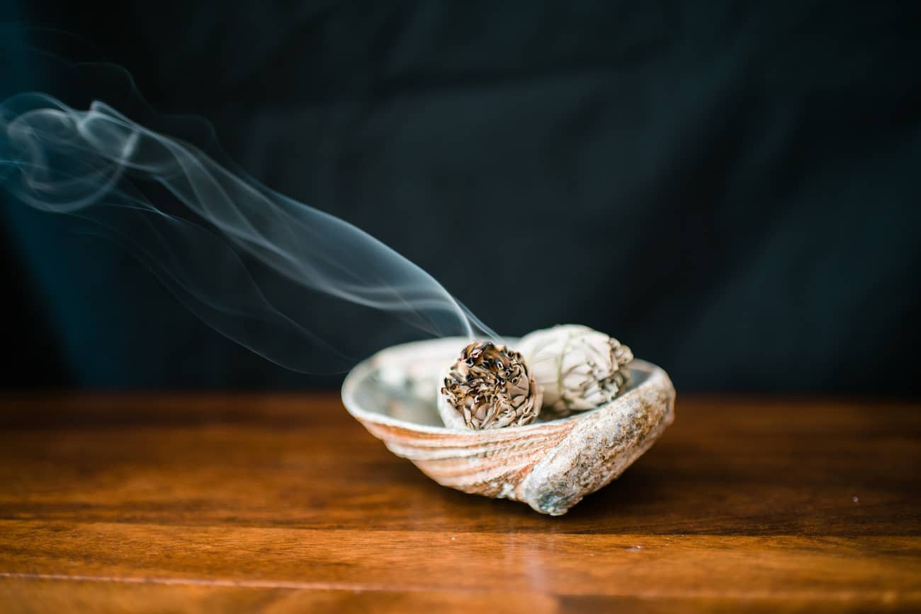 how to smudge your home using a homemade smudging stick in a bowl lightly lit with smoke coming out, all on a wooden table