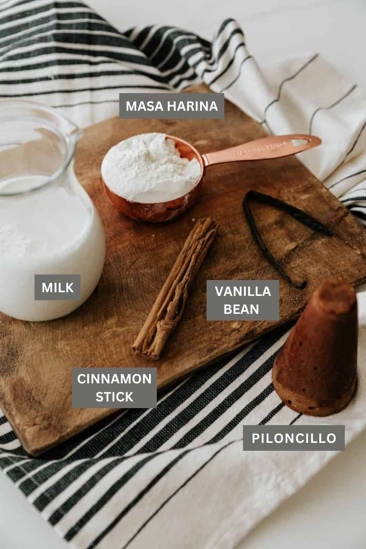 milk, cinnamon stick, vanilla bean, measuring cup of masa harina and a cone of piloncillo for making atole de maiz.