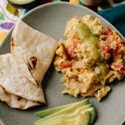 migas on a grey plate with avocado slices and toasted flour tortillas for making breakfast tacos.