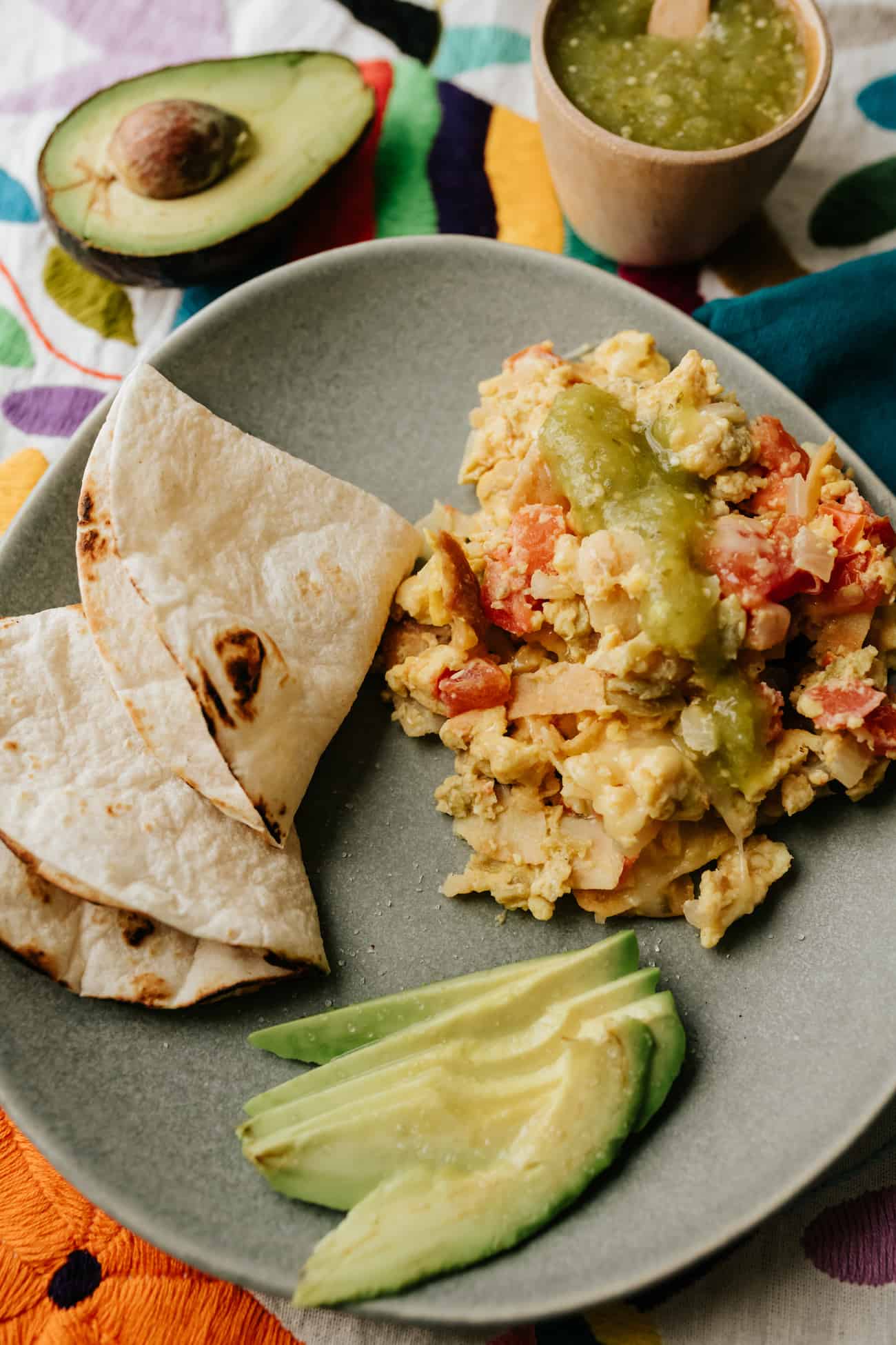 migas with a side of flour tortillas for making breakfast tacos on a gray round plate with slices of avocado and a side of salsa verde.