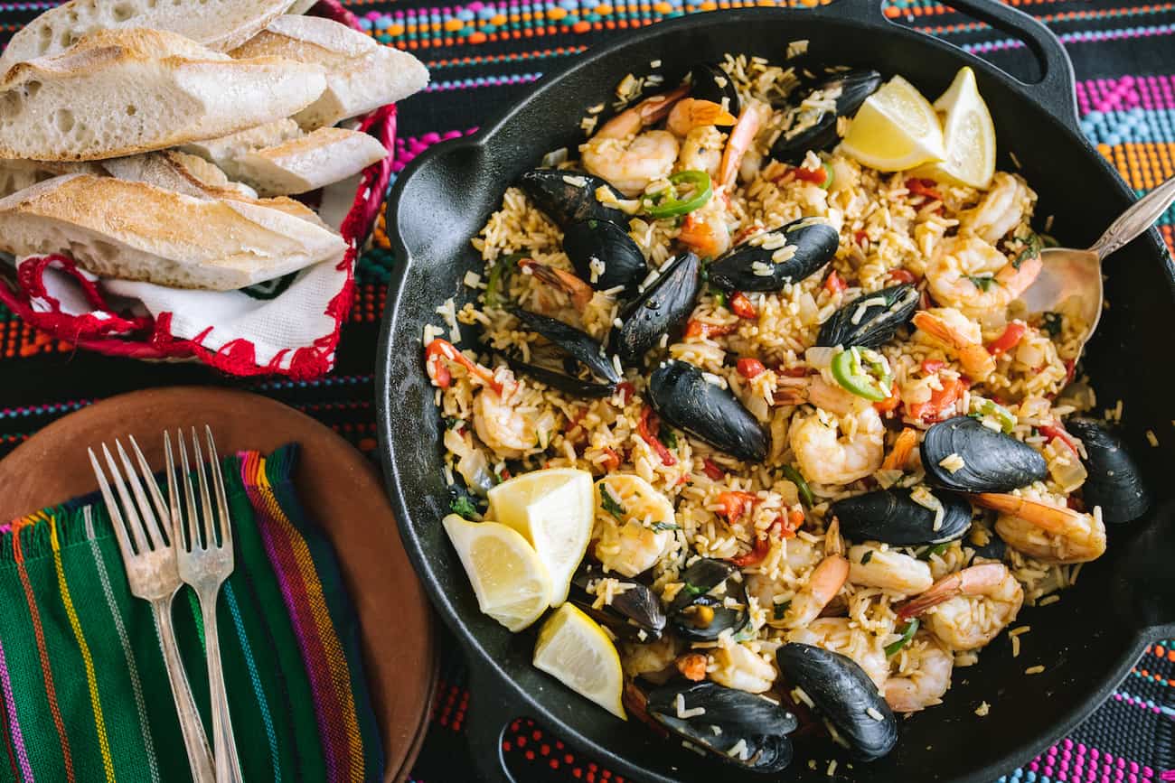 overhead shot of mussel and shrimp paella de marisco in a cast iron pan with a bowl of baguette slices nearby.
