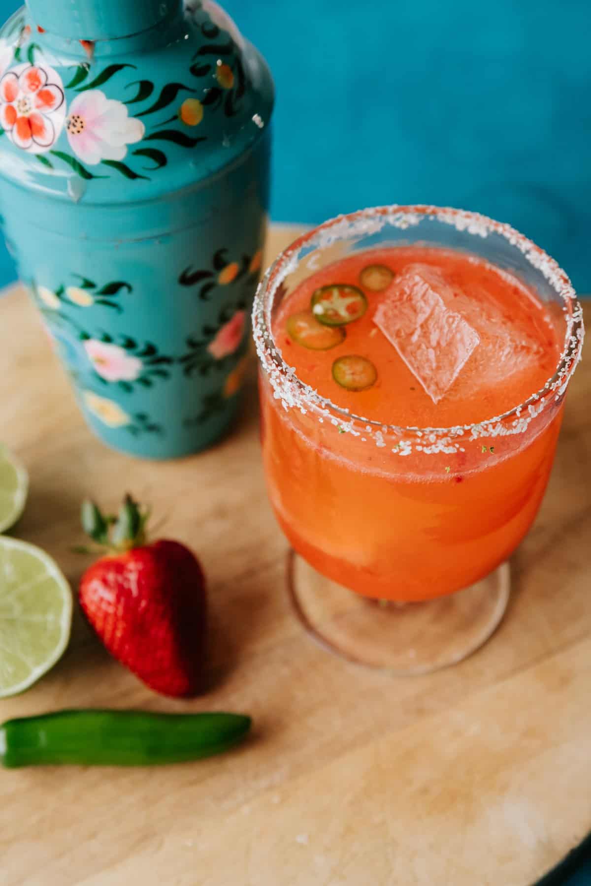 Strawberry jalapeño margarita on the rocks in a clear footed glass with a turquoise floral cocktail shaker in the background.