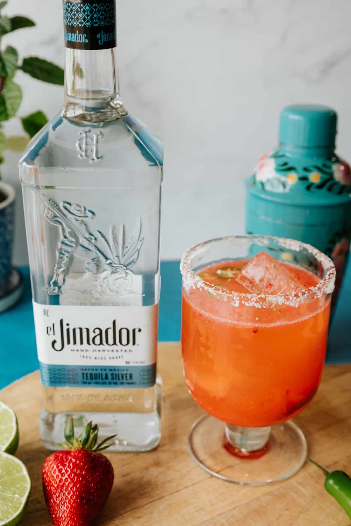 Strawberry jalapeño margarita in a clear footed glass with a salt and lime zest rim next to a bottle of el jimador tequila silver.