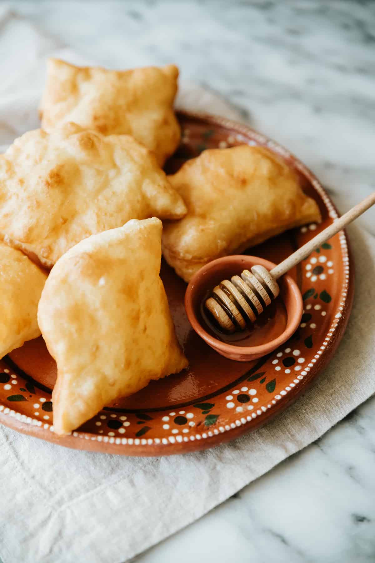 45 degree angle shot of plain New Mexican sopaipillas on a terracotta plate with a small ramekin of honey with a wooden honey dipper in it. 