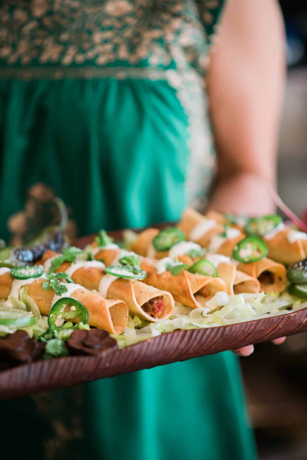 Latina in a green dress holding a platter of Mexican flautas on a wooden tray with a bed of shredded lettuce and drizzled with crema, sliced jalapenos, fresh cilantro, and a sprinkling of cotija cheese.