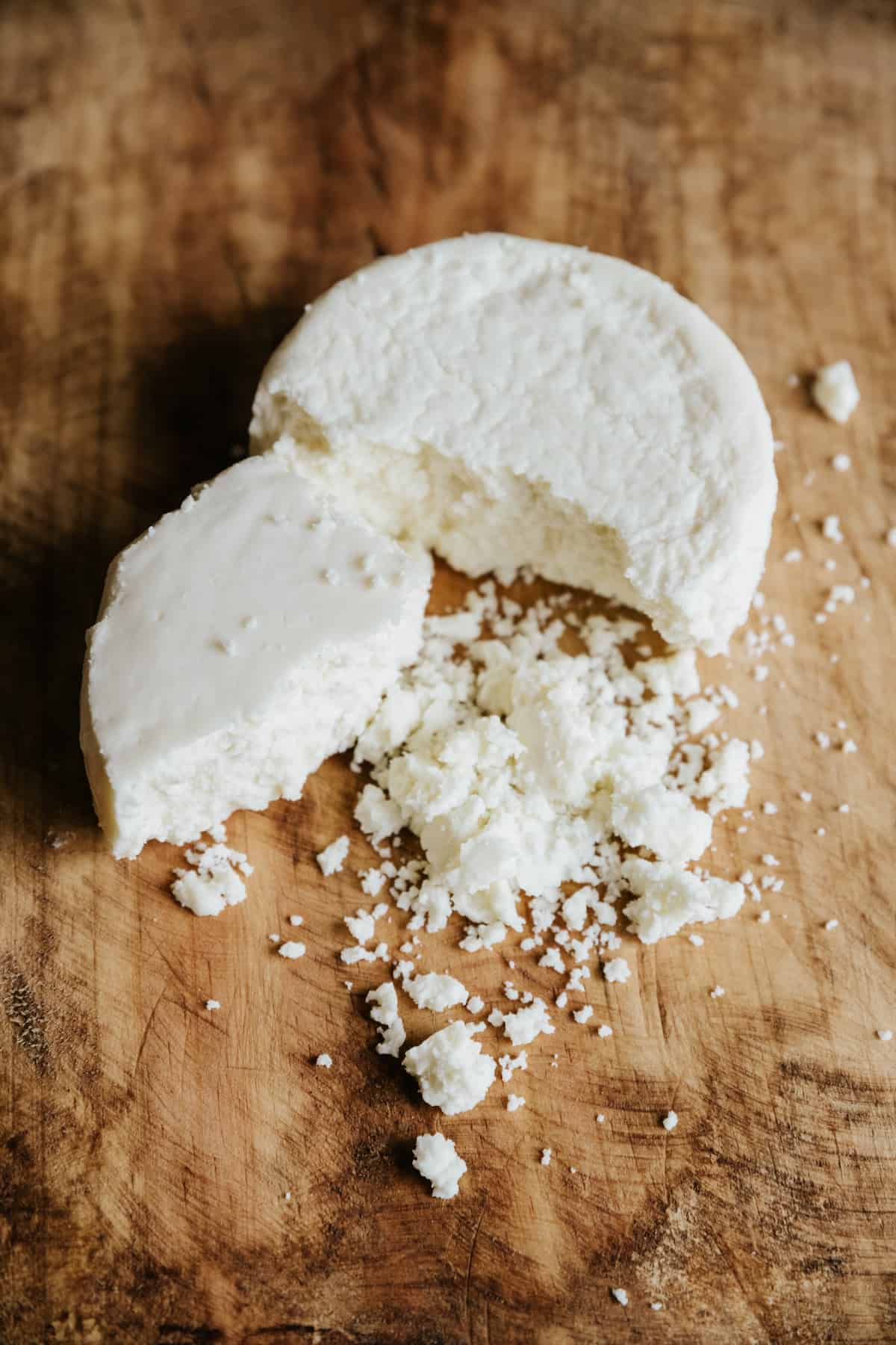 Crumbled cotija cheese on a wooden cutting board, with some of the round of queso still intact.