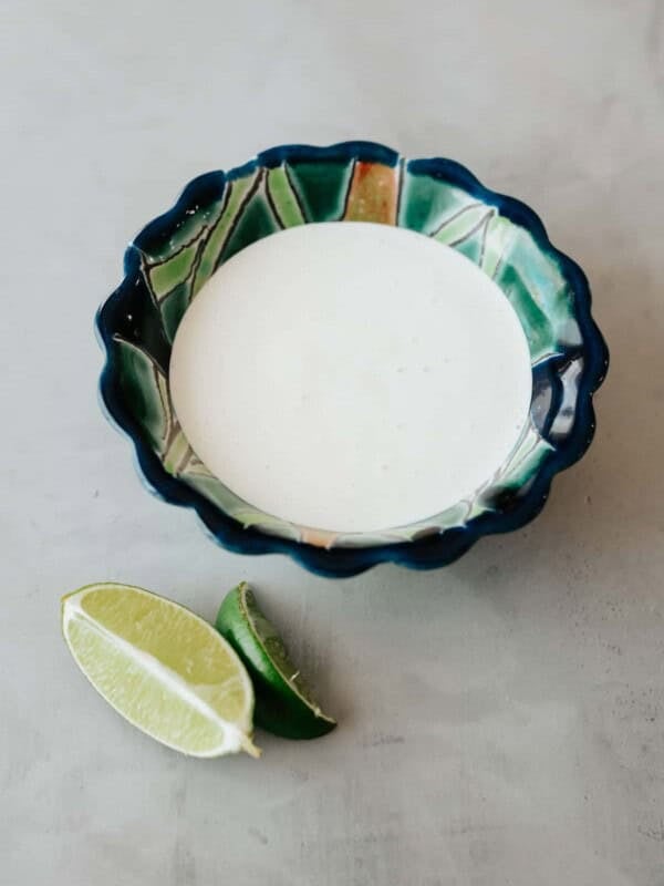 blue and green painted scalloped bowl filled with homemade crema mexicana on a grey table with a few fresh lime wedges to the side.