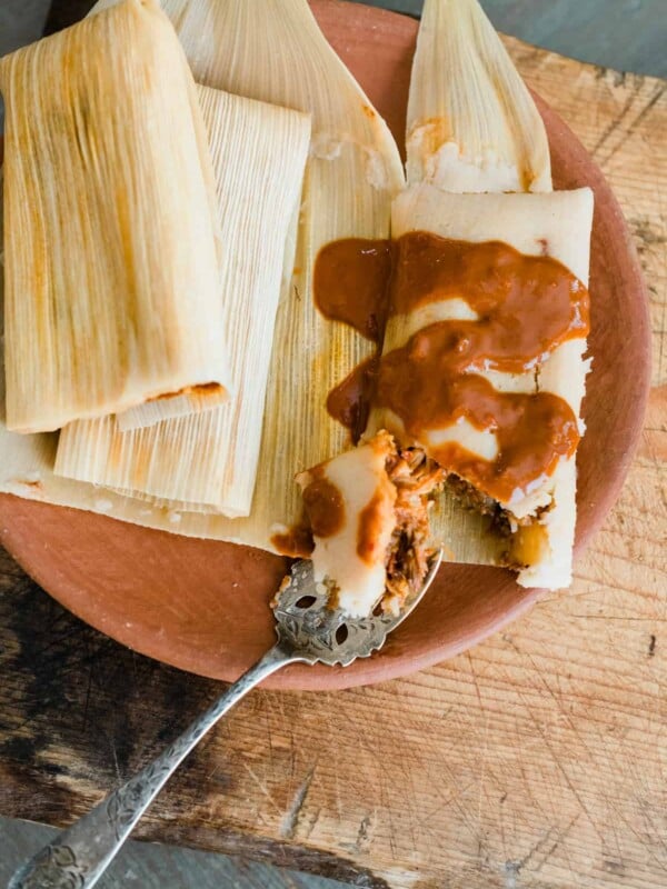 two wrapped chicken mole tamales and one unwrapped tamale doused in extra mole sauce on an earthenware plate.