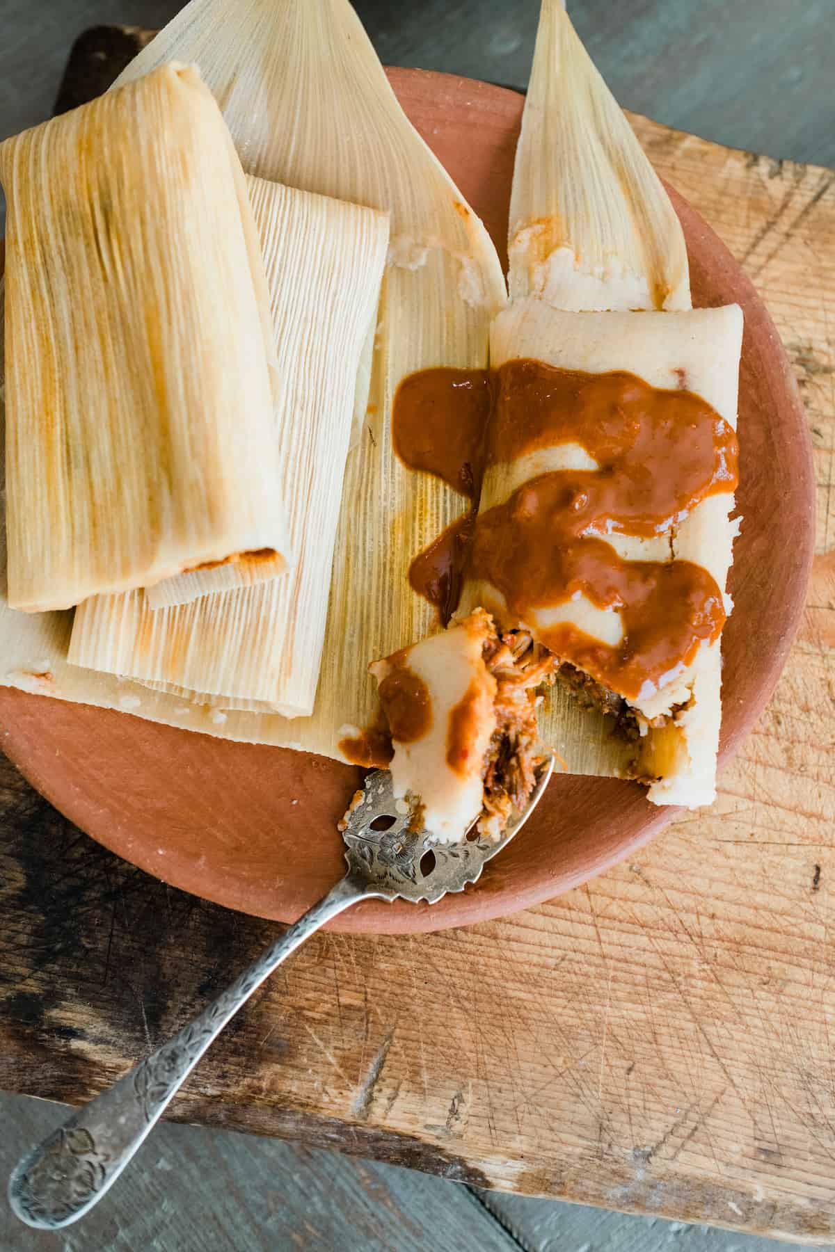 two wrapped chicken mole tamales and one unwrapped tamale doused in extra mole sauce on an earthenware plate. 