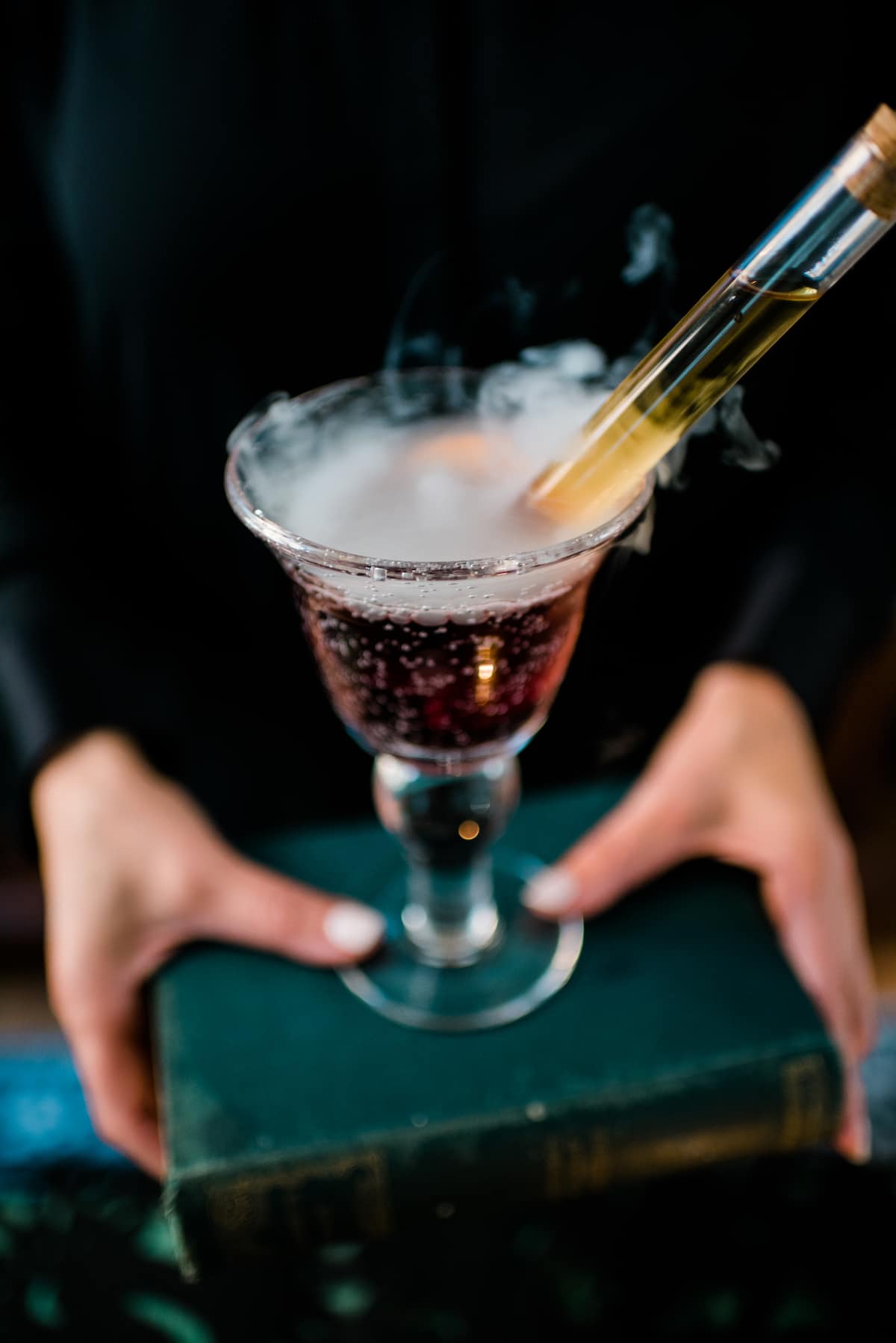 Hands holding a goblet of witch's brew sangria on a green book with dry ice fog spilling over and a test tube of liquor sticking out of it.