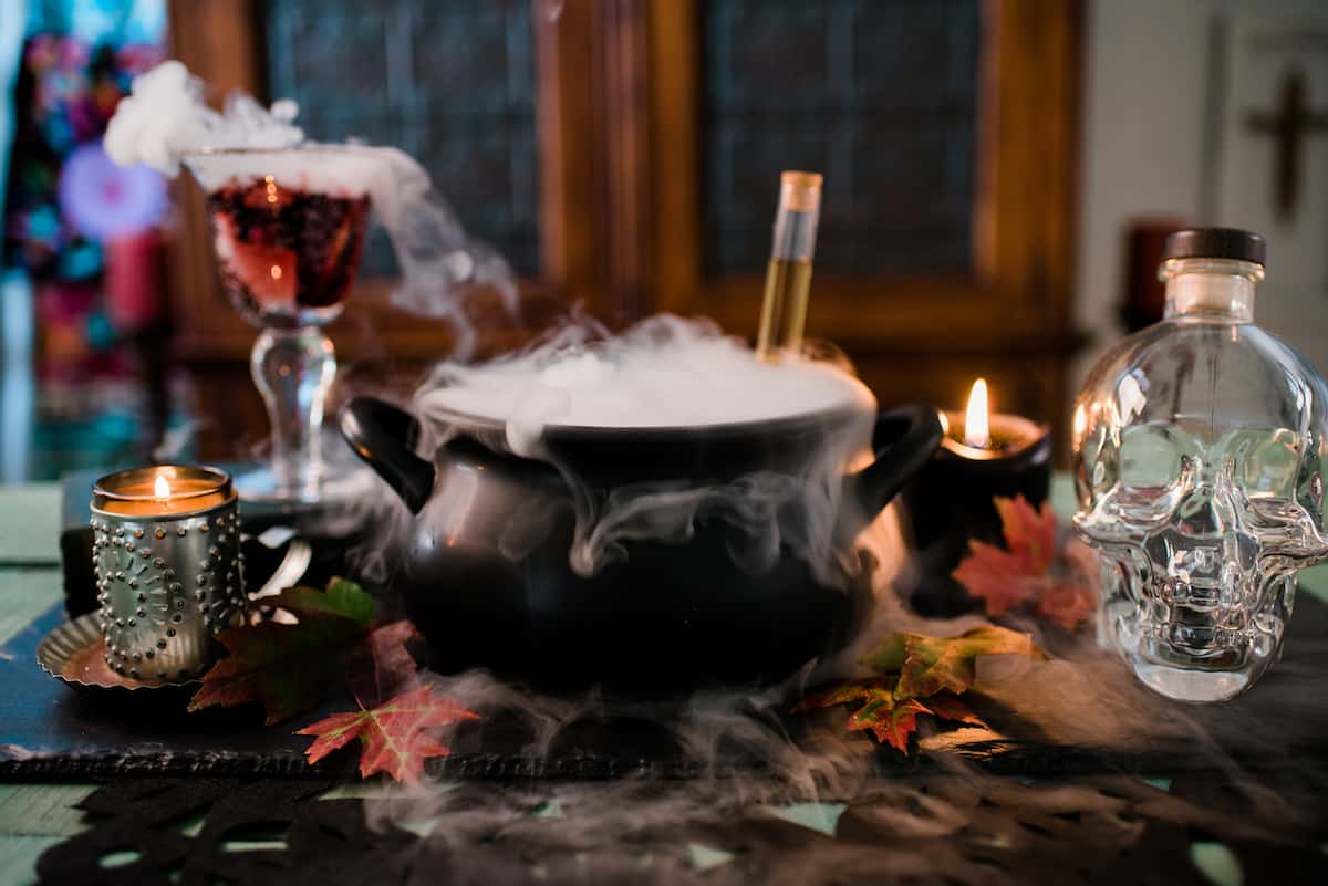 black witches cauldron overflowing with fog from dry ice next to a lit black candle, a bottle of liquor in the shape of a skull, and a goblet of sangria.