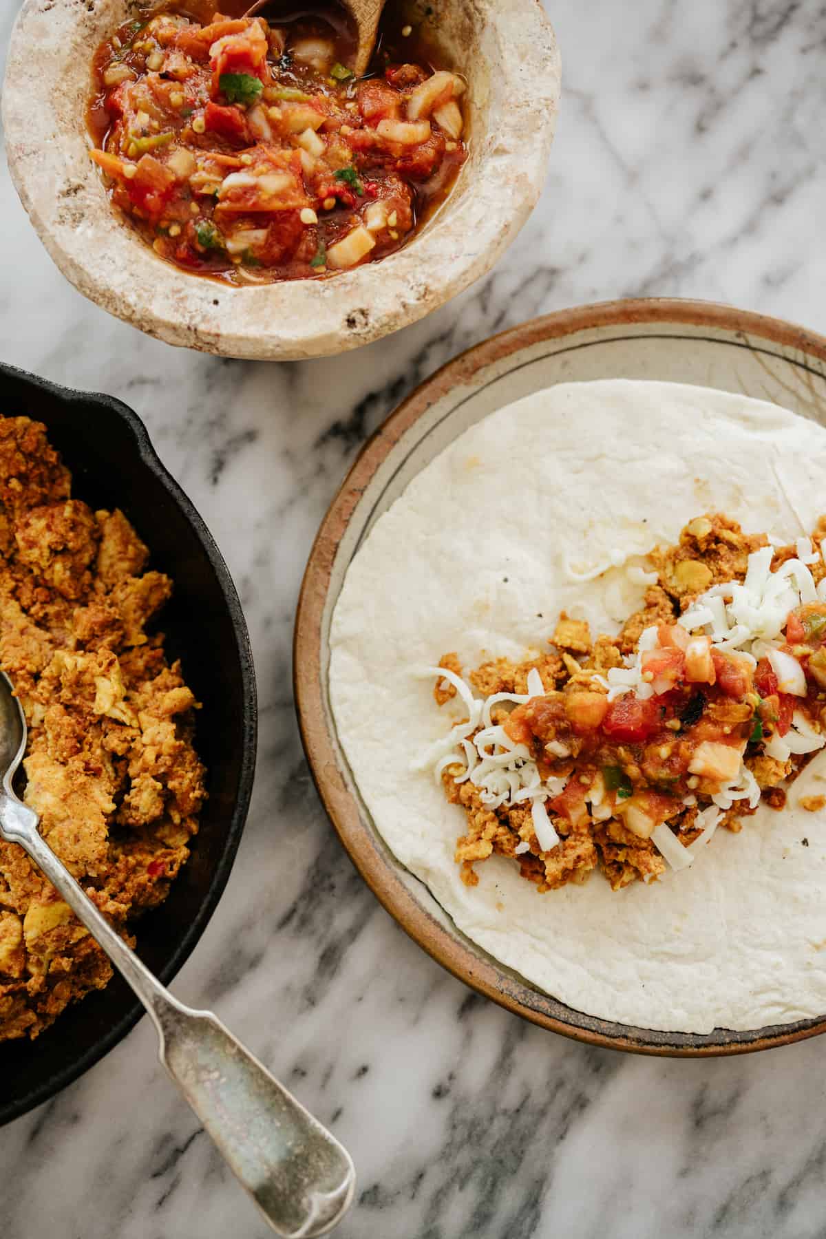 shot showing chorizo and egg breakfast burrito assembly — a plate with an open-faced tortilla layered with breakfast burrito ingredients next to a cast iron pan of the egg and chorizo mix and a bowl of salsa.