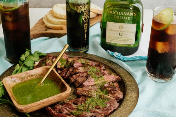 bucola fernet, whisky and cola cocktail on a table with a serving platter of argentinian skirt steak with chimichurri sauce..