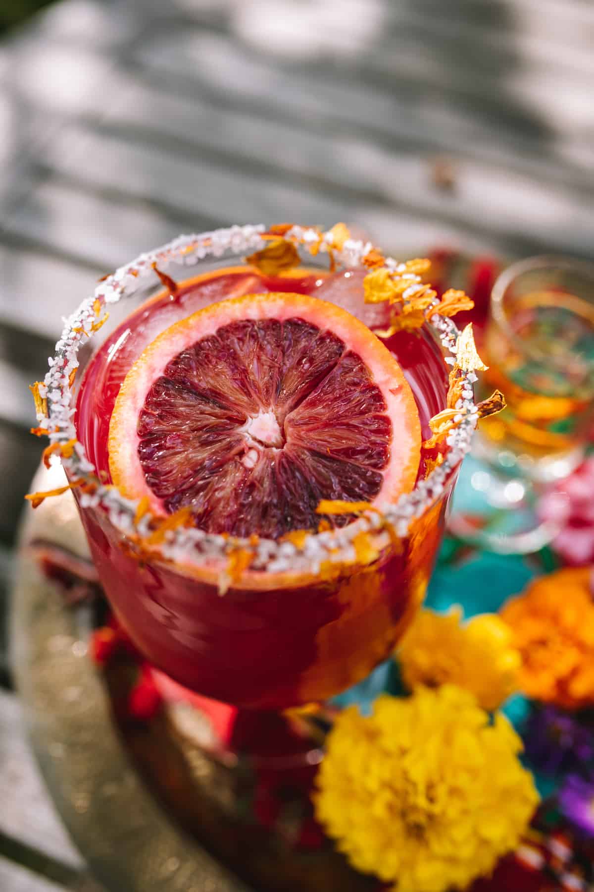 close up image of a blood orange margarita in a glass with a salt and marigold petal rim and a slice of blood orange as garnish.