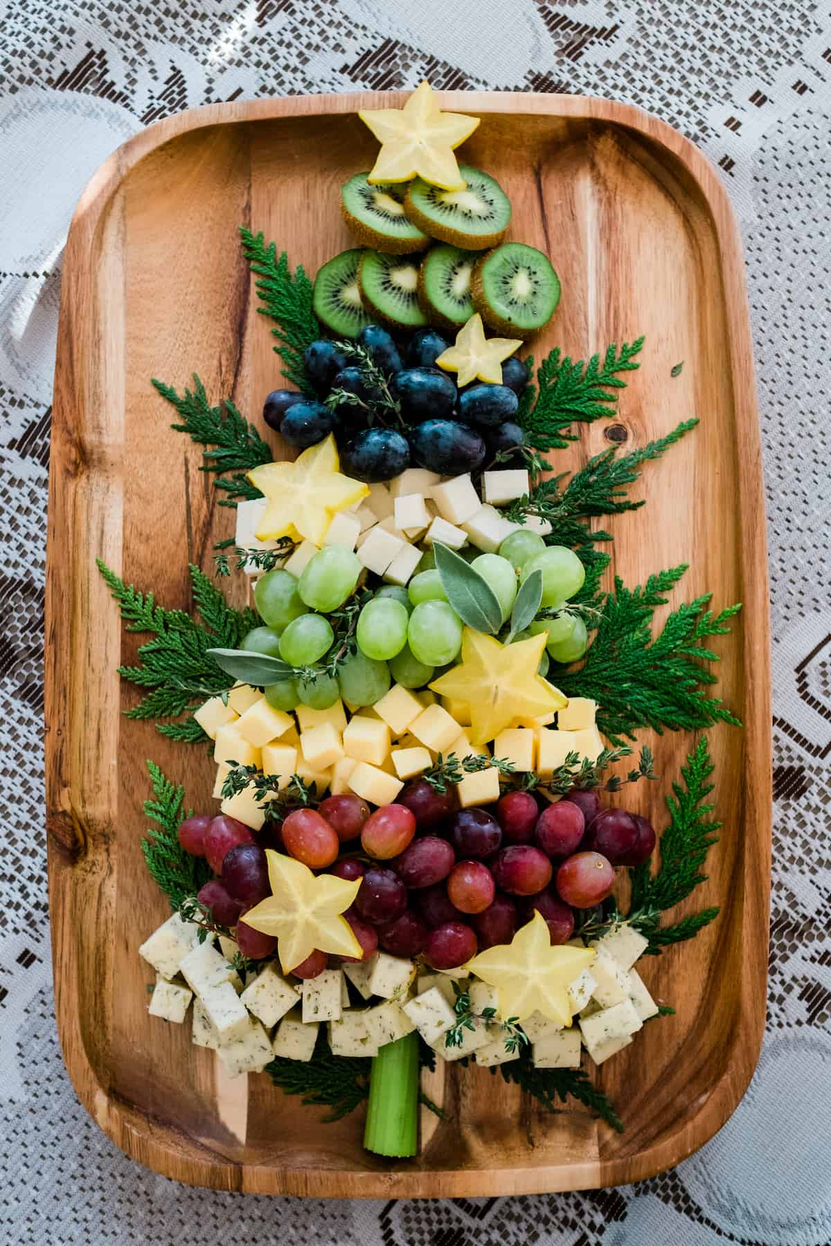 Christmas tree shaped cheese platter on a wooden tray kiwi grapes star fruit and cheese surrounded by evergreen branches. 