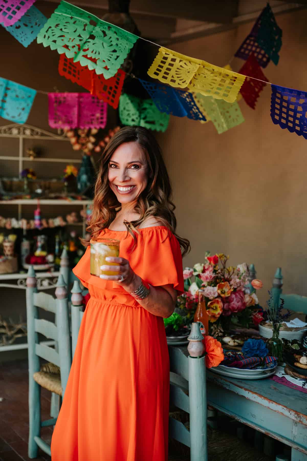 Yvette Marquez-Sharpnack author of Muy Bueno standing in front of a table holding a margarita with Papel picado hanging above ready for a fiesta. 