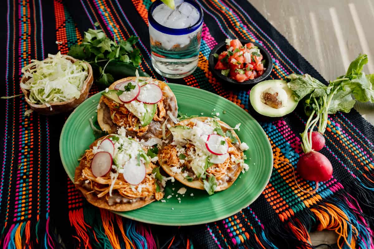 green serving plate with 3 chicken tinga tostadas on a colorful woven placemat, a blue rimmed glass of water, a bowl of shredded lettuce, a molcajete filled with pico de gallo, a halved avocado, 2 radishes with their stems attached, and a bunch of cilantro.