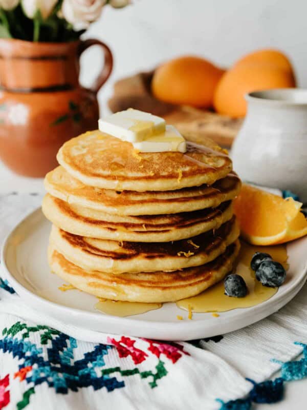 stack of 4 orange buttermilk pancakes on a white plate topped with maple syrup and fresh blueberries with a wedge of orange.