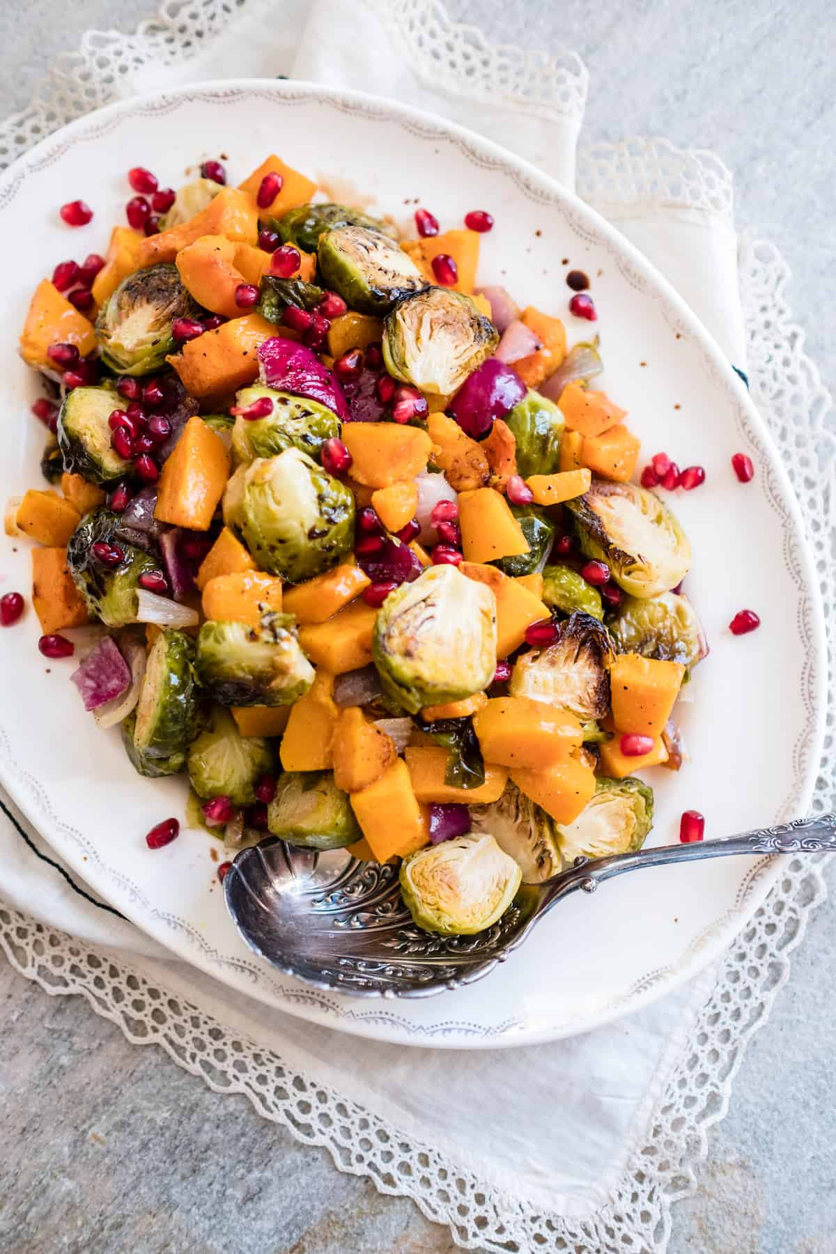overhead shot of an oval white serving platter filled with roasted butternut squash and burssels sprouts with red onion, pomegranate arils and a drizzle of pomegranate molasses with a silver serving spoon. 