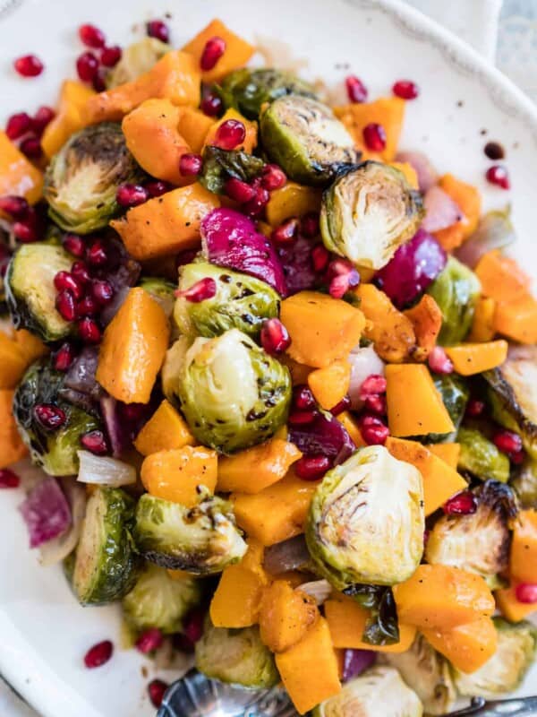 Serving platter of roasted butternut squash, burssels sprouts, red onions, and pomegranate seeds with a silver serving spoon.