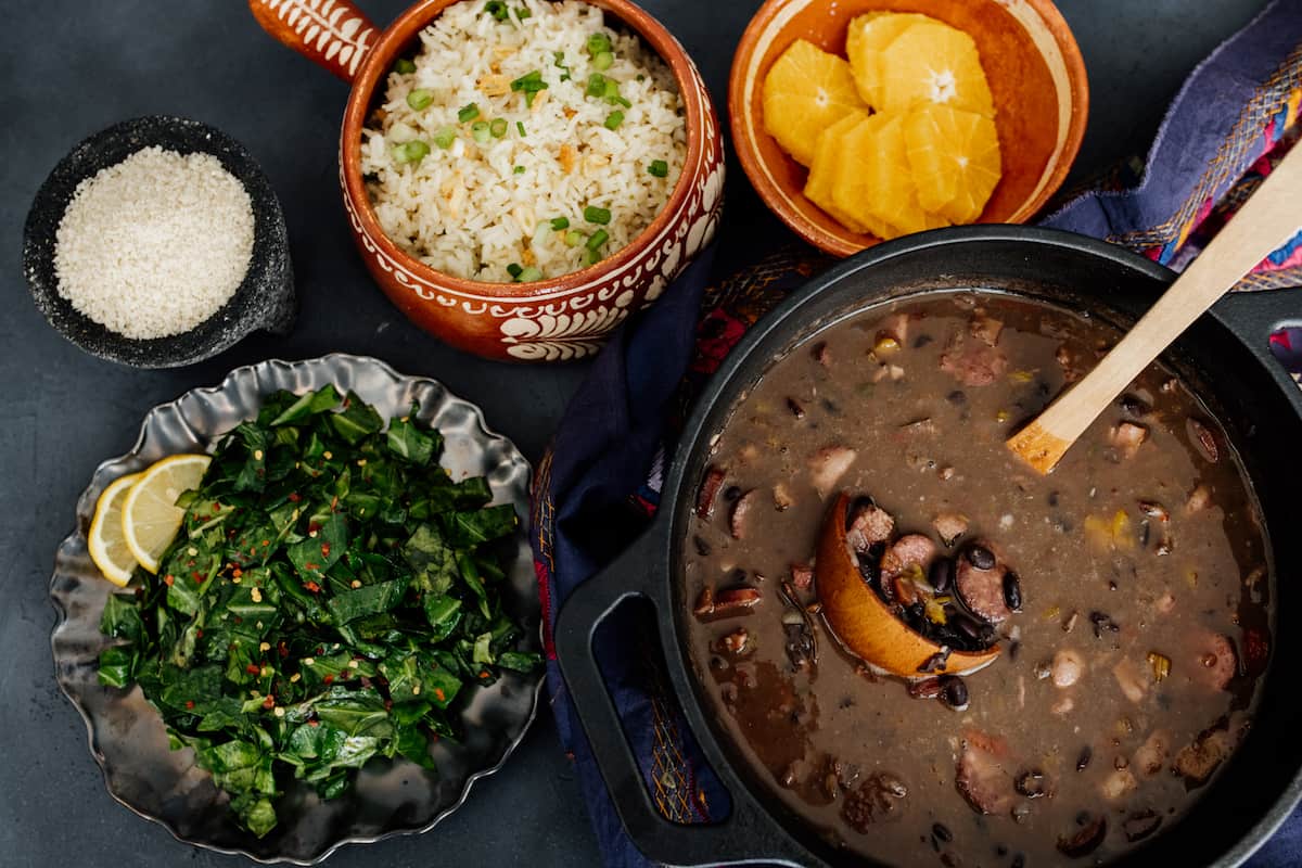 horizontal hero shot of a pot of Brazilian feijoada stew next to a serving plate of collard greens, a plate of orange slices, and a bowl of garlic butter rice.