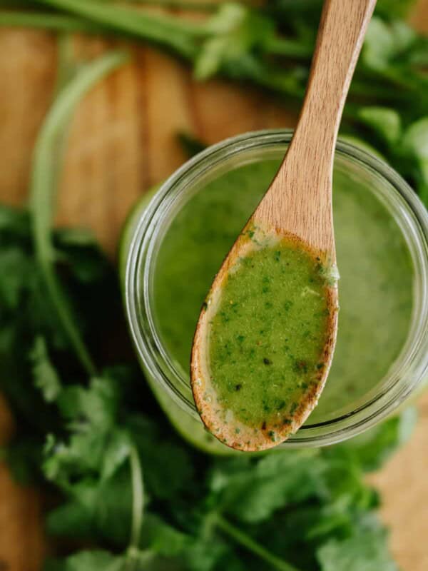 overhead shot of a glass mason jar filled with parsley cilantro chimichurri sauce with a wooden spoonful of it in focus and the jar and fresh herbs out of focus below it.