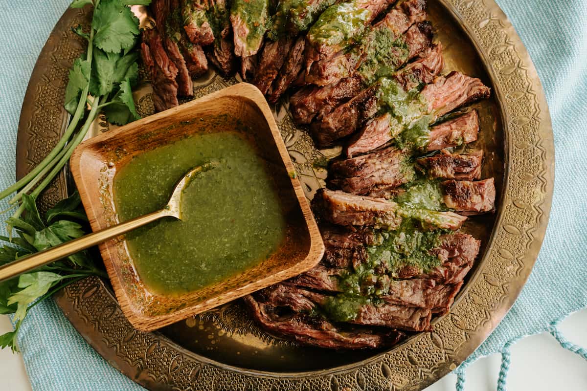 round brass serving platter with sliced flank steak and a square wooden bowl of parsley cilantro chimichurri drizzled over the top.