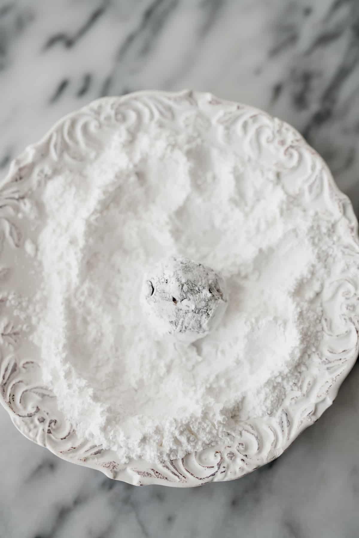 white plate with powdered sugar and a crinkle cookie dough ball that has been coated in powdered sugar.