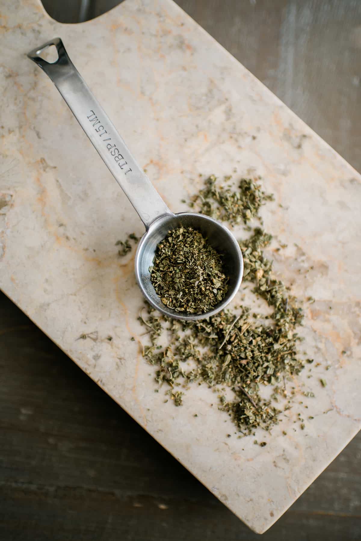 silver tablespoon measure with dried oregano on a marble surface.