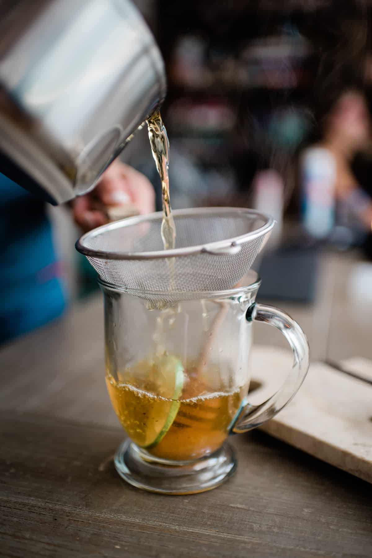 showing how to strain oregano tea from a saucepan through a mesh strainer into a mug.