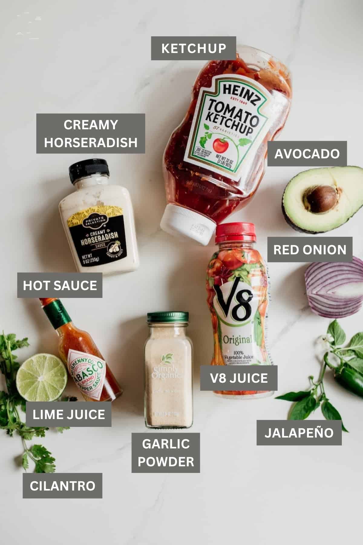 Ingredients for Coctel de Camarones (Mexican Shrimp Cocktail) including creamy horseradish, ketchup, V8 vegetable juice, hot sauce, garlic powder, red onion, avocado, lime, cilantro, and jalapeño on a white surface