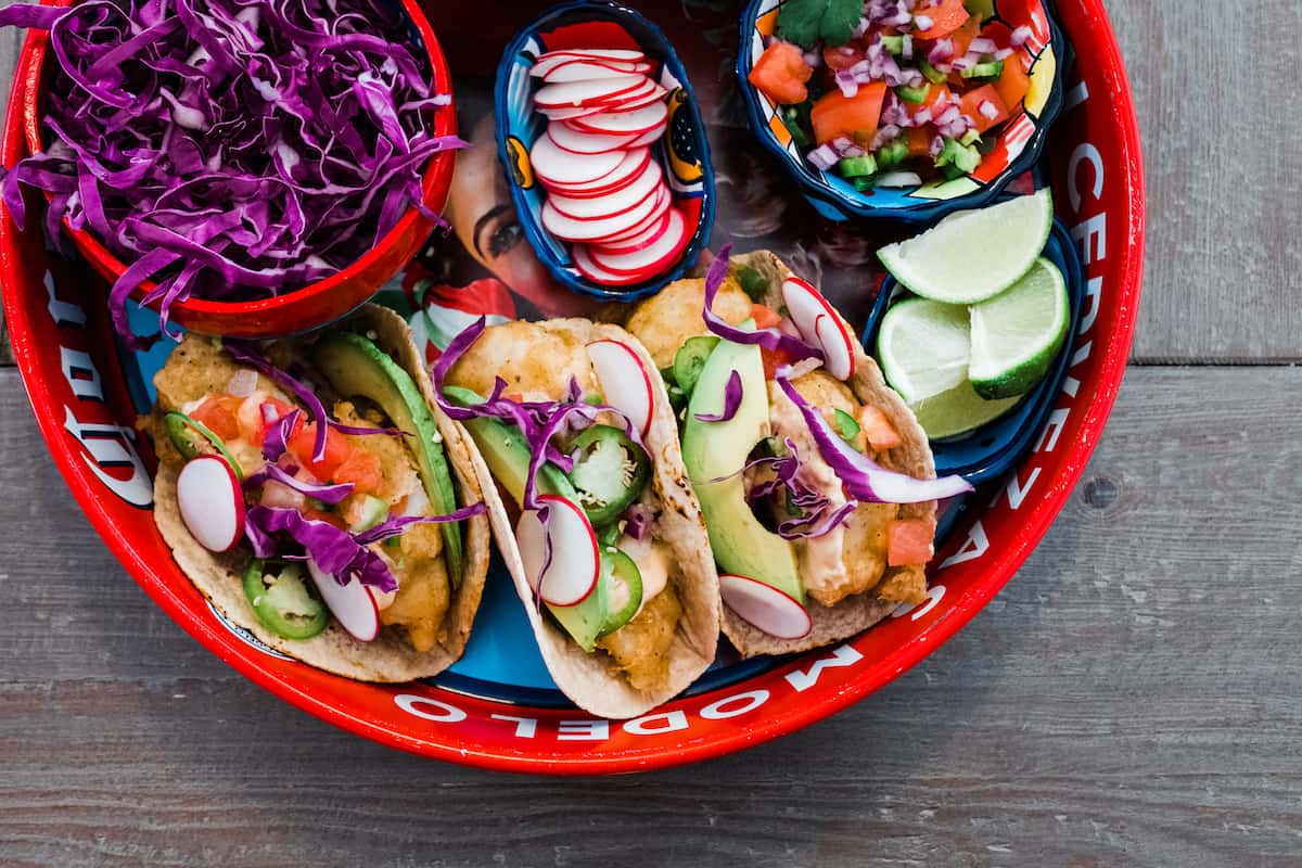 Beer-Battered Fish Tacos with all the toppings and garnishes in a Mexican tray on a gray table.