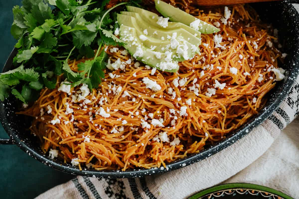 45 degree angle shot of an oval black earthenware serving dish filled with fideo seco and topped with crumbled queso fresco, a handful of cilantro leaves, and sliced avocado. 