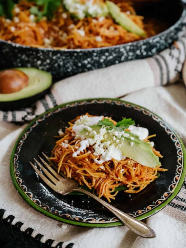 45 degree angle shot of a black plate with a green rim filled with a serving of fideo seco topped with avocado slices, a drizzle of crema, and a sprinkle of queso fresco.