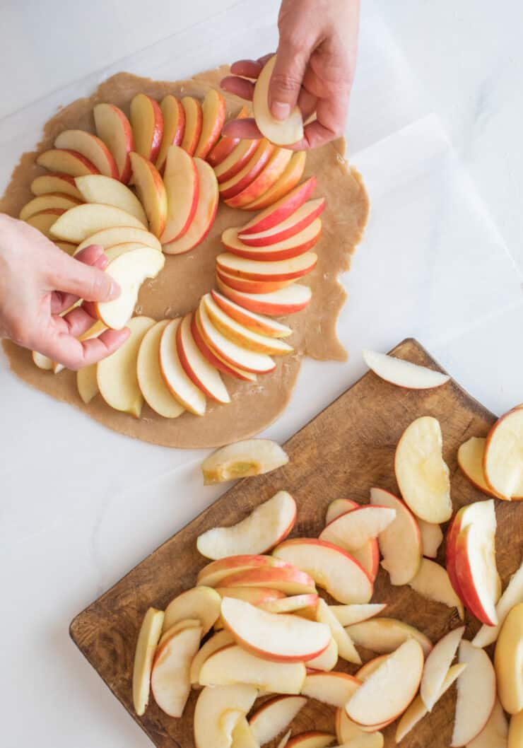 Hands arranging fresh apple slices on rolled-out galette dough.