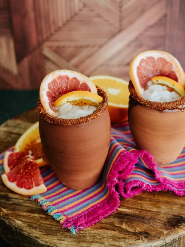 two earthenware cantarito cups rimmed with tajin, filled with ice and skinny cantaritos mix and garnished with orange and grapefruit slices.