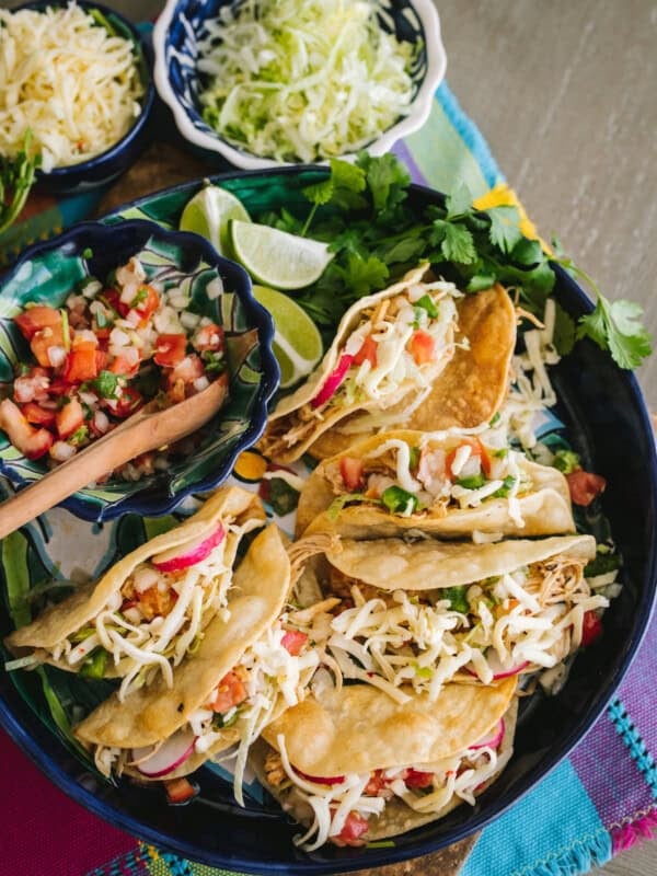 overhead shot of a serving platter of homemade crispy chicken tacos with small serving bowls of toppings like shredded lettuce, shredded cheese, and pico de gallo.