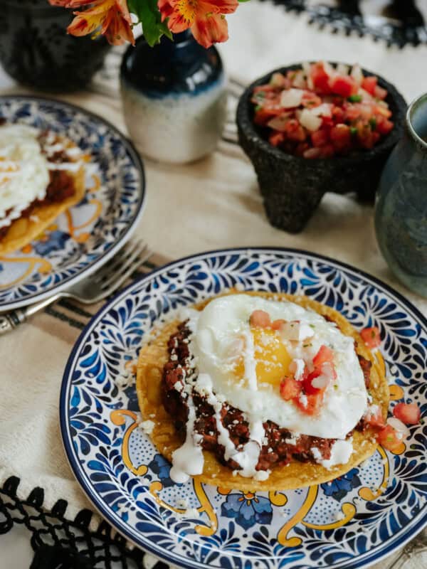 overhead shot of a breakfast table with yellow, blue and white hand-painted plates filled with black bean and chorizo huevos rancheros, cafe de olla, and a molcajete of pico de gallo.