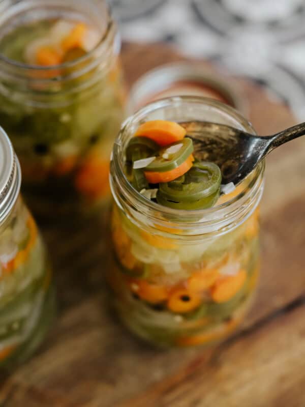 multiple mason jars filled with Mexican escabeche on a wooden cutting board with the closest one with a spoon in it fishing out a serving.