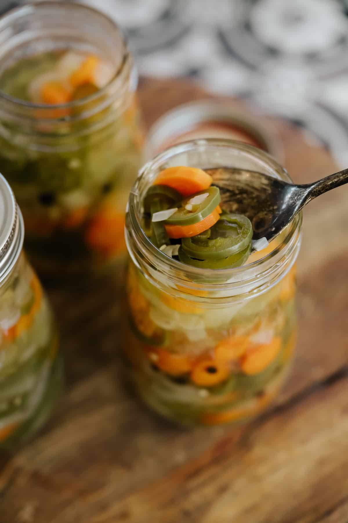 multiple mason jars filled with Mexican escabeche on a wooden cutting board with the closest one with a spoon in it fishing out a serving.