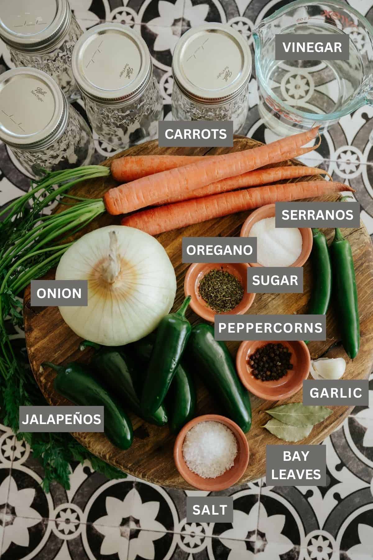 Carrots and chile peppers with spices needed to make Mexican escabeche laid out on a wooden cutting board next to sanitized mason jars for canning. 