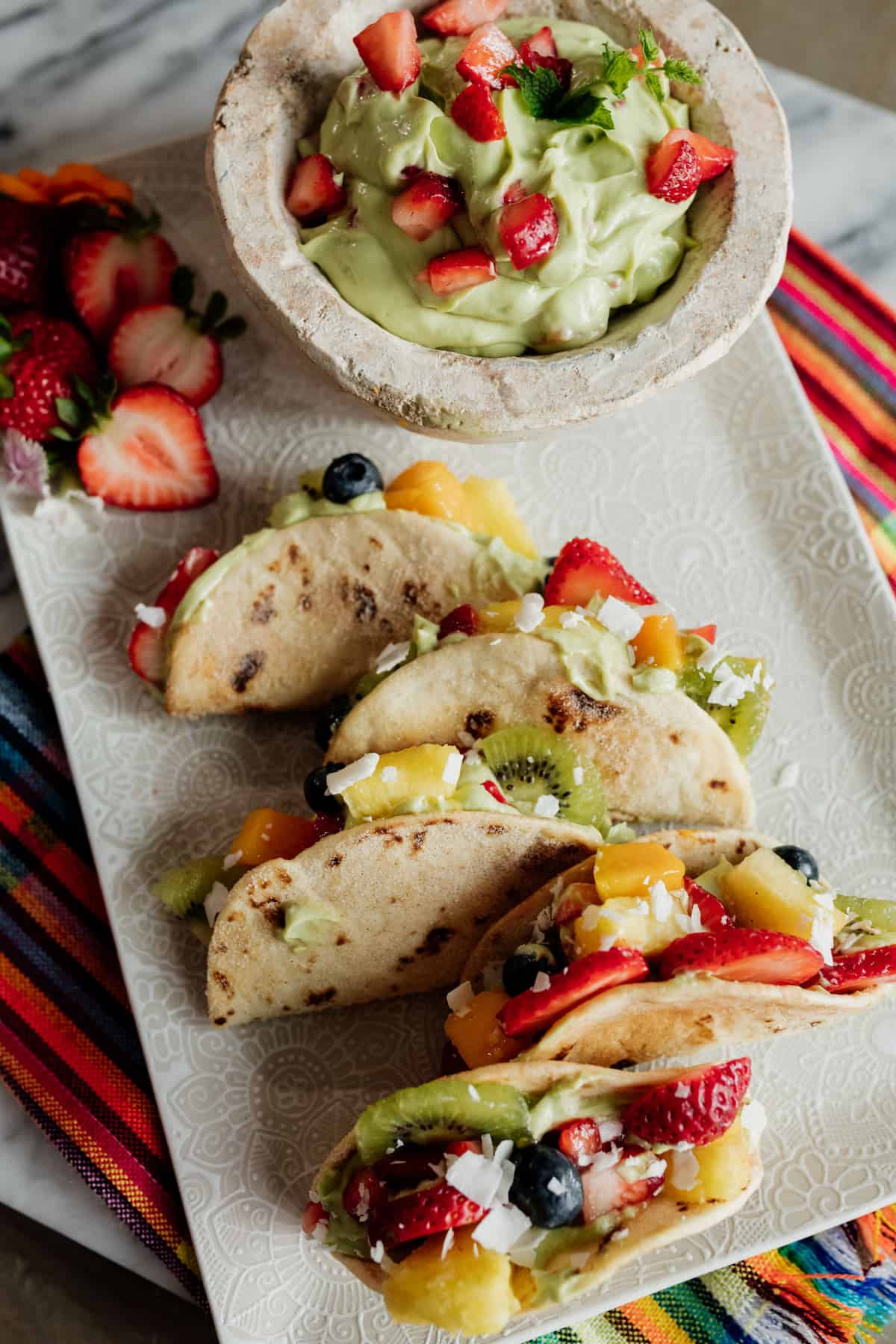 dessert tacos filled with avocado yogurt cheesecake filling and sliced fruits and berries on a white platter.