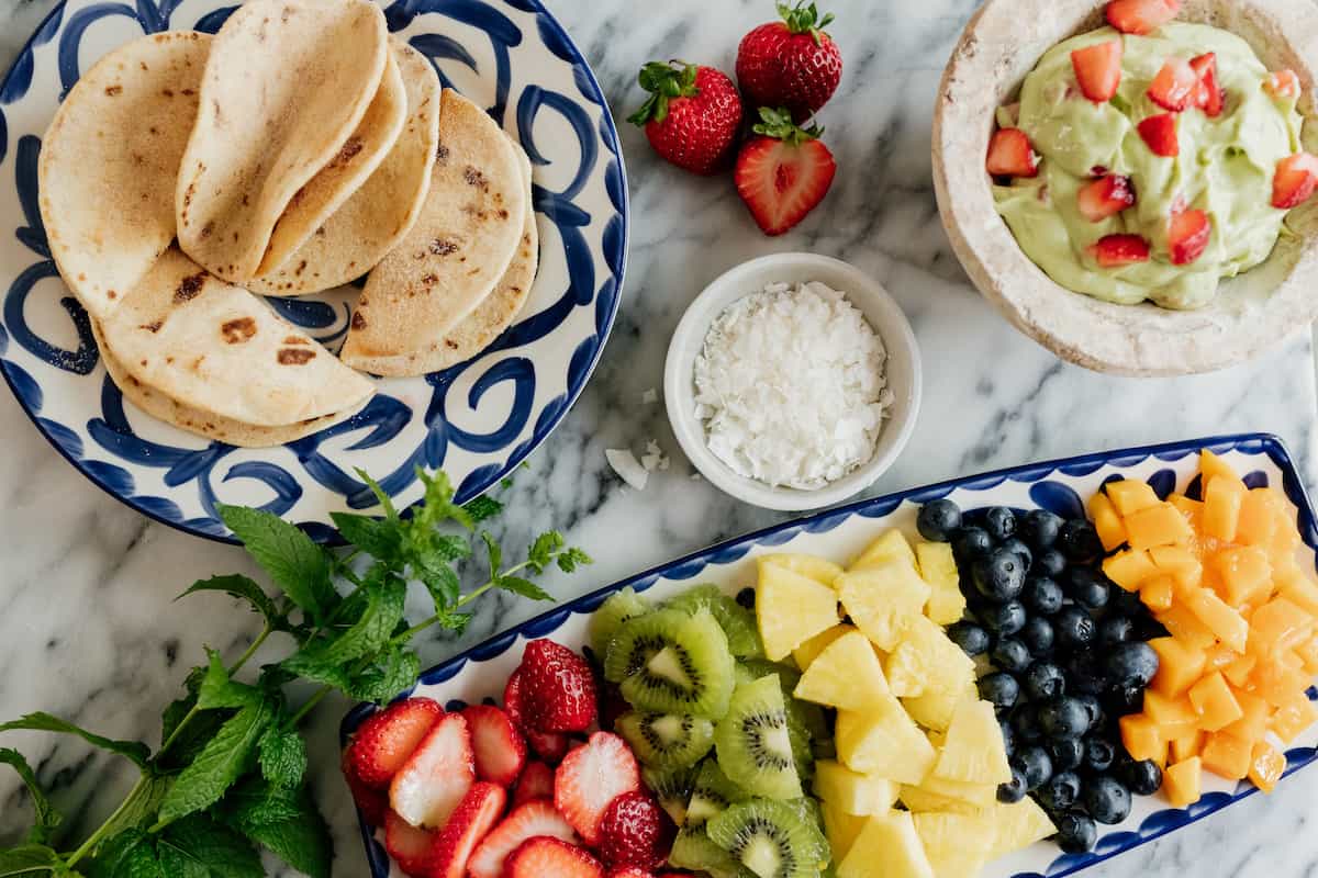 dessert taco buffet with cinnamon sugar taco shells on one plate, an assortment of cut fresh fruit on another plate, a molcajete filled with avocado yogurt cheesecake filling, and a small bowl of shredded coconut.