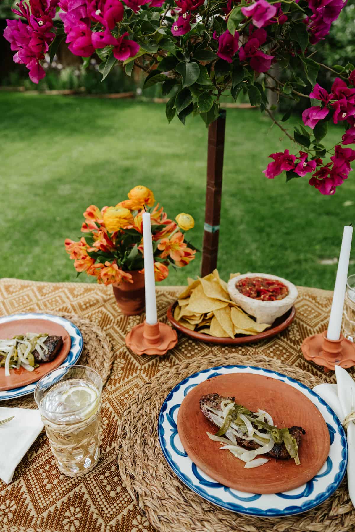 neutral colored dinner table under a pergola with bougainvillea crawling on it with tampiqueña steaks and a molcajete with fresh salsa. 